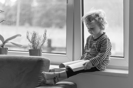 Young caucasian boy with curly hair reading book and sitting on windowsill.の写真素材