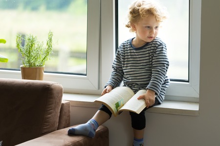 Young caucasian boy with curly hair reading book and sitting on windowsill.