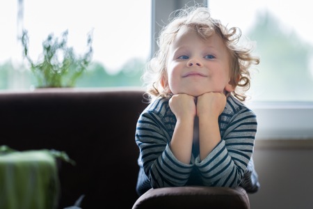 Young boy with blonde curly hair posing. Boy propels his head with his hands and lying on armchair.の写真素材