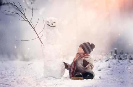 Young boy sitting near snowman in winter scenery. Winter landscape with snowman and small boy.の写真素材