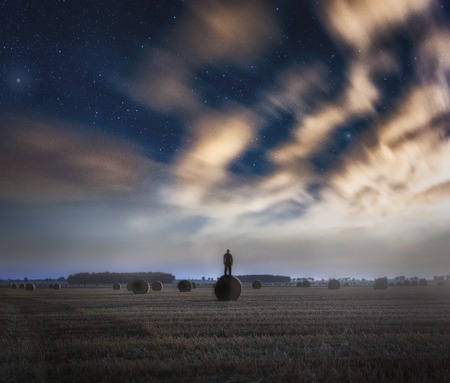 Strange man standing on field on straw bale under starry sky. Fine art landscape.の写真素材