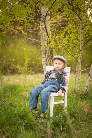 Young blonde boy relaxing on white old chair standing under birch tree in outdoor.の写真素材