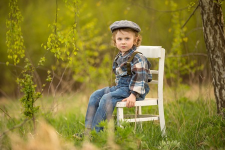 Young blonde boy relaxing on white old chair standing under birch tree in outdoor.の写真素材