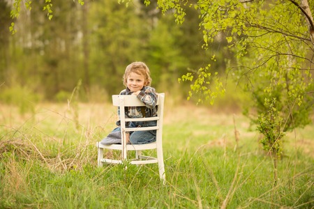 Young blonde boy relaxing on white old chair standing under birch tree in outdoor.の写真素材