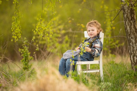 Young blonde boy relaxing on white old chair standing under birch tree in outdoor.の写真素材