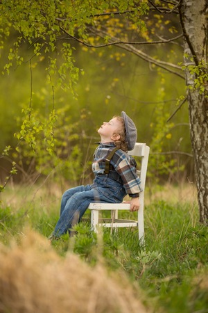 Young blonde boy relaxing on white old chair standing under birch tree in outdoor.の写真素材