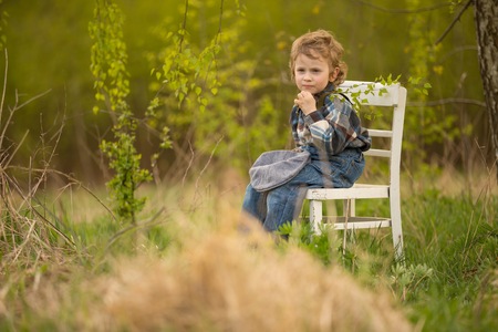 Young blonde boy relaxing on white old chair standing under birch tree in outdoor.の写真素材