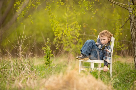 Young blonde boy relaxing on white old chair standing under birch tree in outdoor.の写真素材