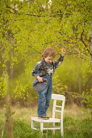 Young blonde boy standing on white old chair under birch tree in outdoor.の写真素材