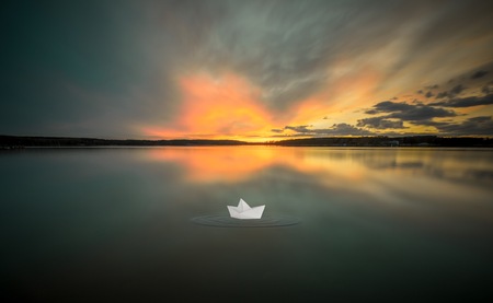 Dreamy surreal landscape with paper boat on lake water and sunset sky reflected in water.