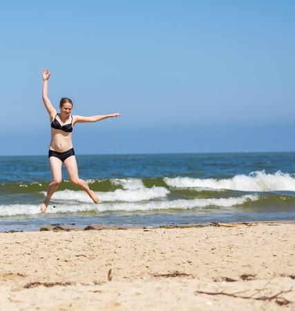 Happy young woman jumping on sea shore at sunny summer day. Blonde woman enjoys the holidays.の写真素材