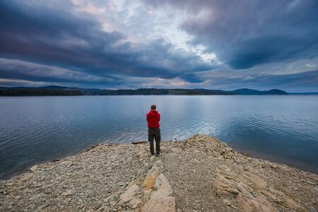 Man standing on lake shore under dark sky. Solinskie lake in Bieszczady mountainsの写真素材