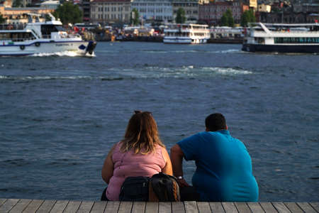 Istanbul, Turkey - July 13, 2019: An obese young couple is sitting and watching the scenery at The Golden Horn, Istanbul. There are tour boats are carrying passengers.のeditorial素材