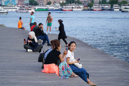 Istanbul, Turkey - July 13, 2019: Chinese young women are resting at coast of The Golden Horn. Also they have smart phones on their hands and some young muslim women using cell phones at the back.のeditorial素材