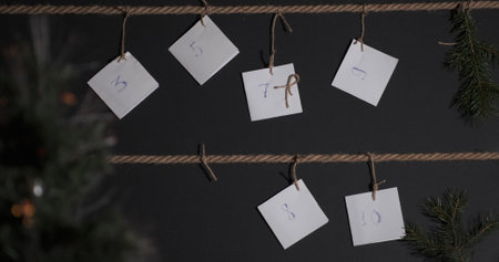homemade DIY advent calendar, created on a black background and tied with a craft string, in the foreground is a Christmas tree with a garland, male hands tie another tagの写真素材