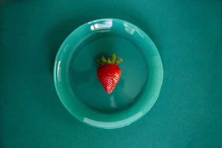 Top view of one red big juicy strawberry in the center of a turquoise glossy plate on a turquoise background with copy spaceの写真素材