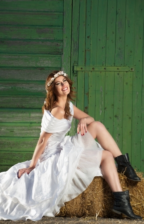 Smiling bride in wedingdress and boots. She is happy and sitting on a haystack against a green barn doorの写真素材