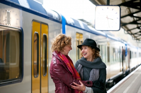 Two mature women friends welcoming each other with a smile and a warm embrace at an outdoor train station platformの写真素材