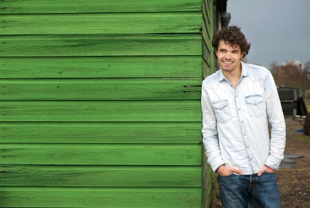 Happy male model leaning against green wooden wall outdoor. Hands in pockets and smile expression on his faceの写真素材