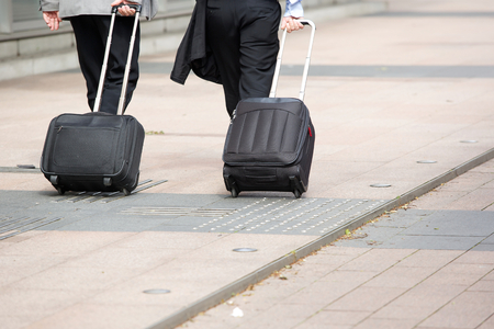 Two businessmen walking with wheeled luggage on city sidewalkの写真素材