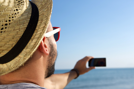 Portrait from behind of a young man taking a selfie with cell phone at the seaの写真素材