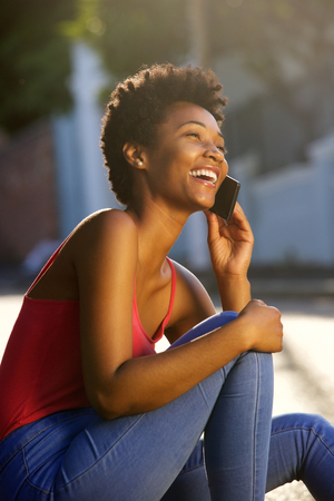 Portrait of happy young woman sitting along a street and talking on cell phoneの写真素材