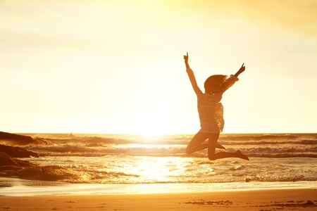 Silhouette portrait of young woman jumping for joy at beach during sunsetの写真素材