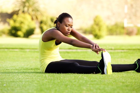 Portrait of young african woman sitting on grass stretching to reach toesの写真素材
