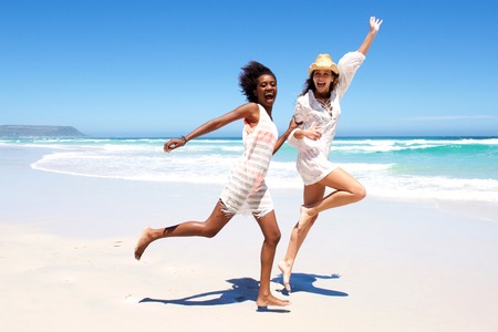 Full body portrait of two young women friends laughing and running on the beachの写真素材