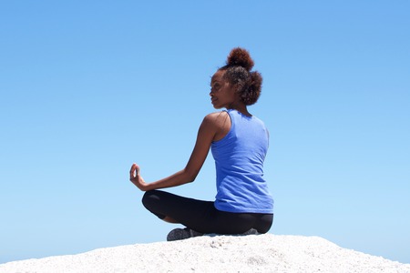 Rear view portrait of young african woman sitting at beach in yoga poseの写真素材