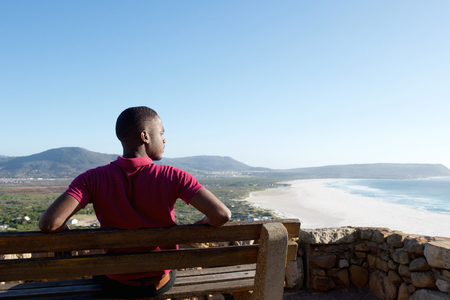 Rear view portrait of young african man sitting relaxed on a bench and looking at the sea, man on a holiday.の写真素材