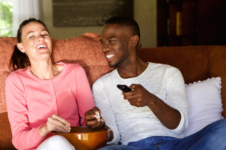 Portrait of happy young couple sitting on sofa watching movie and eating popcorn at homeの写真素材