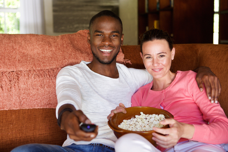 Portrait of mixed race young couple sitting on living room sofa watching a movie and eating popcornの写真素材