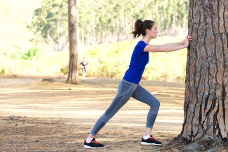 Full length side portrait of sporty woman stretching against tree before workout