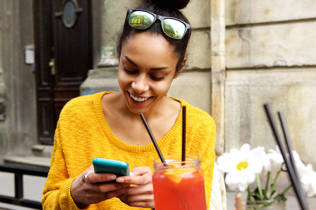 Close up portrait of happy young african woman sitting at outdoor cafe using mobile phone and...