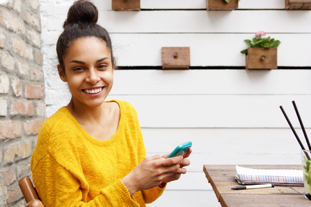 Close up portrait of cheerful young black woman sitting at cafe with mobile phoneの写真素材