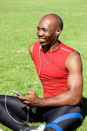 Portrait of african sports man sitting on grass outdoors and listening to music on headphonesの写真素材