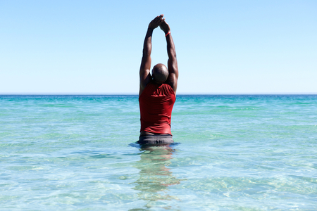 Rear portrait of fit young african man standing in sea and doing arms stretching workoutの写真素材