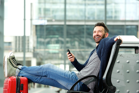 Portrait of happy mature man sitting with travel bag and mobile phone at the train stationの写真素材