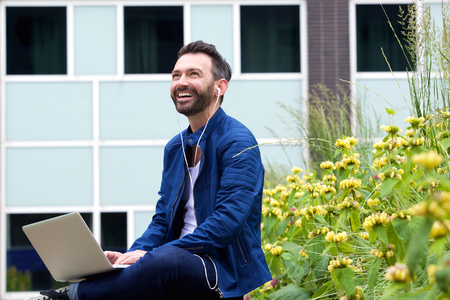 Portrait of happy man wearing earphones and sitting outdoors with laptop, listening to music from laptop.の写真素材