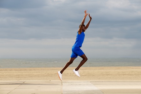 Full body side portrait of fit young african american man jogging at beach with arms raisedの写真素材