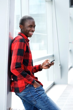 Side portrait of young african man with mobile phone leaning to wall and smilingの写真素材