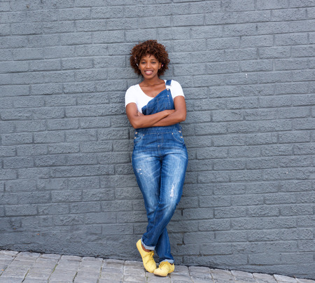 Full length portrait of smiling african woman in dungarees standing against gray wallの写真素材