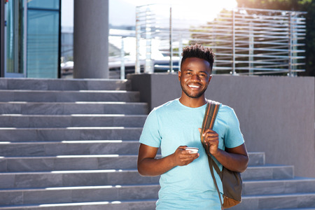 Portrait of smiling african student walking by stairs with bag and smart phone