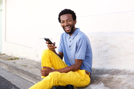 Portrait of laughing man holding smart phone sitting by wallの写真素材