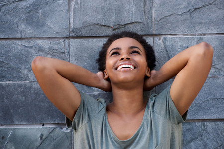 Close up portrait of beautiful young black woman laughing against gray wall with hands behind headの写真素材