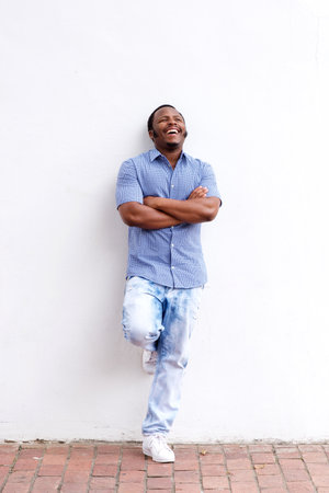 Full body portrait of happy black man leaning against wall and laughingの写真素材