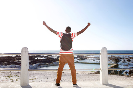 Portrait from behind of young african man traveler standing with arms raised and looking at the beachの写真素材
