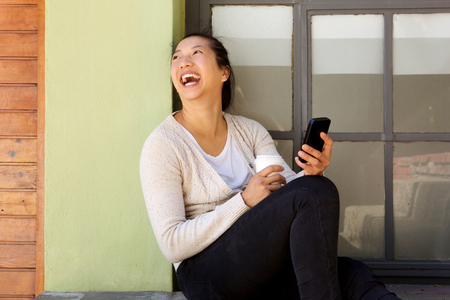 Portrait of happy asian woman sitting outside with a mobile phone looking away and laughingの写真素材
