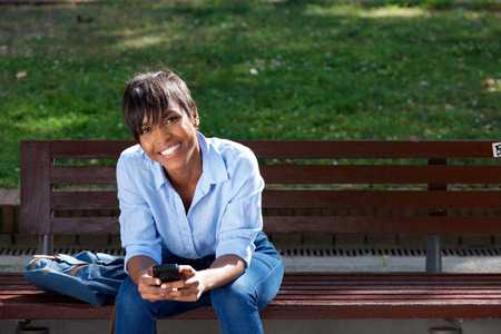 Portrait of attractive young black woman sitting on bench outside with mobile phoneの写真素材